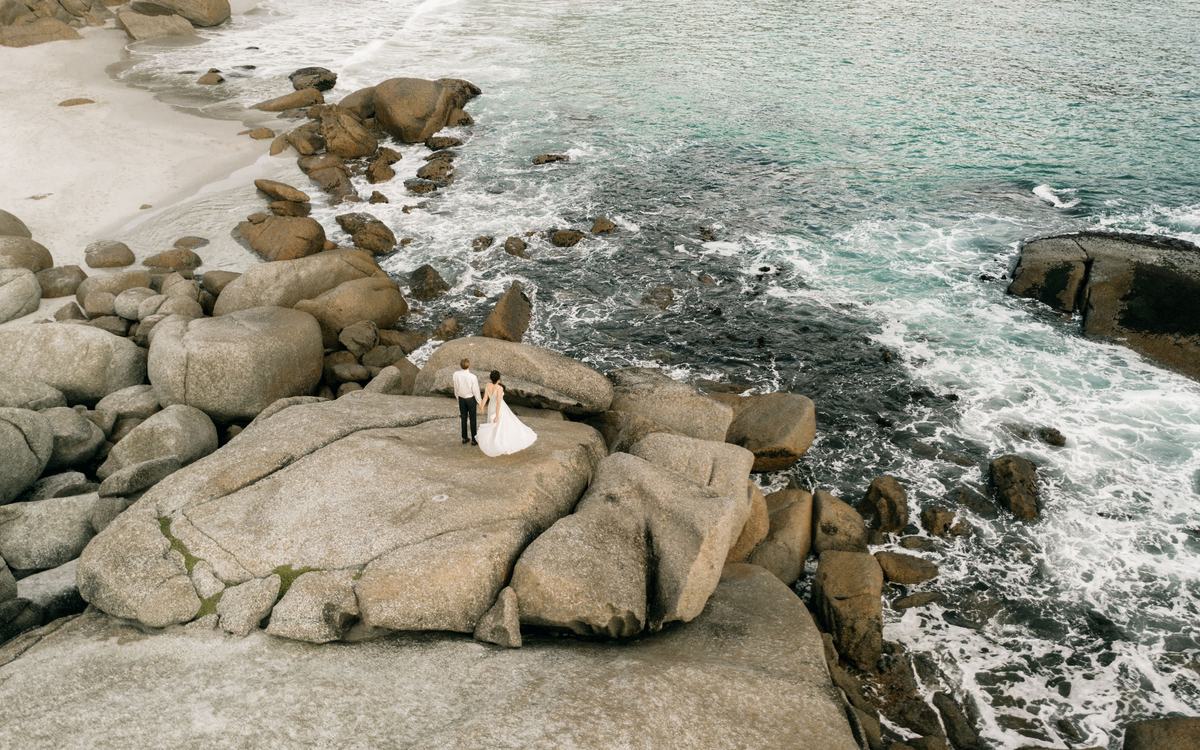Drone view of a bride and groom standing on large granite boulders above a turquoise sea